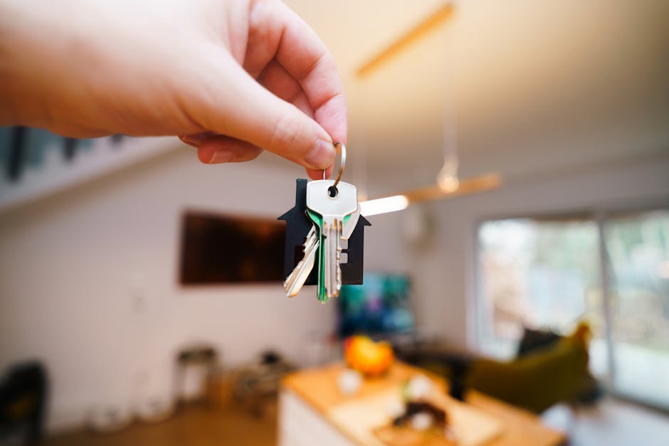 A close-up of a person's hand holding a keyring with several keys, including one with a black plastic head, in a modern living room. The background features a wooden dining table with a bowl of fruit,