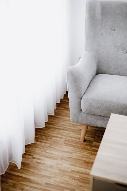 A close-up view of a light grey upholstered armchair positioned in a corner of a room next to sheer white curtains. The armchair has wooden legs and appears clean and well-maintained. The room feature