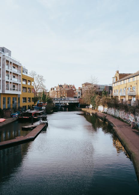 A daytime view of a narrow river with calm, reflective water flowing through an urban area. On the left side, there is a modern, multi-story residential building with a white and yellow facade, featur