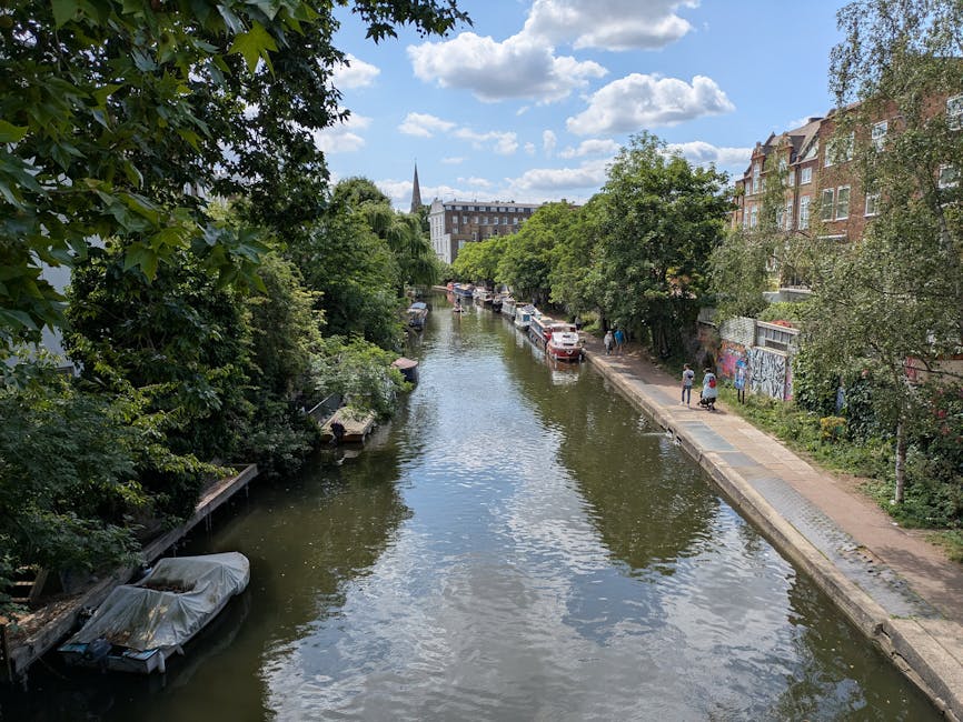 A daytime view of a narrow river with calm, reflective water flowing through an urban area. On the left side, there is a modern, multi-story residential building with a white and yellow facade, featur
